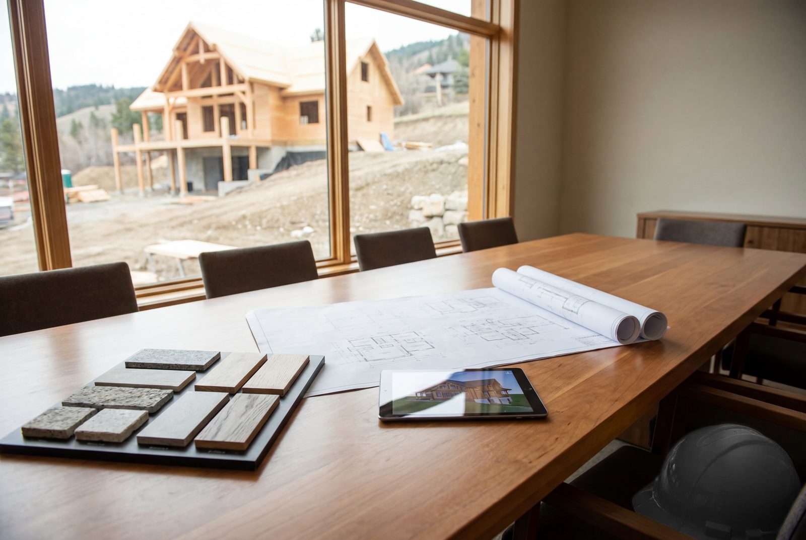 Material samples, drawings, and a rendering tablet on a conference table overlooking an active timber-frame build