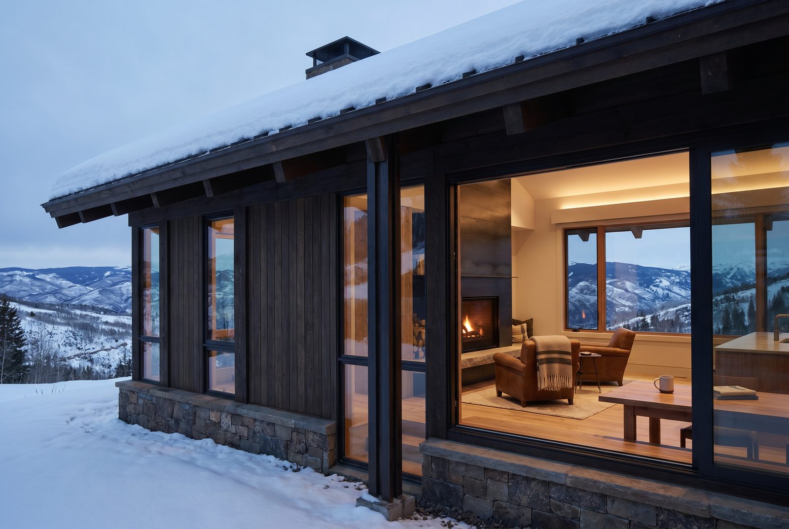 A snow-loaded roof and deep eaves over a warmly lit living room, mountains fading into dusk