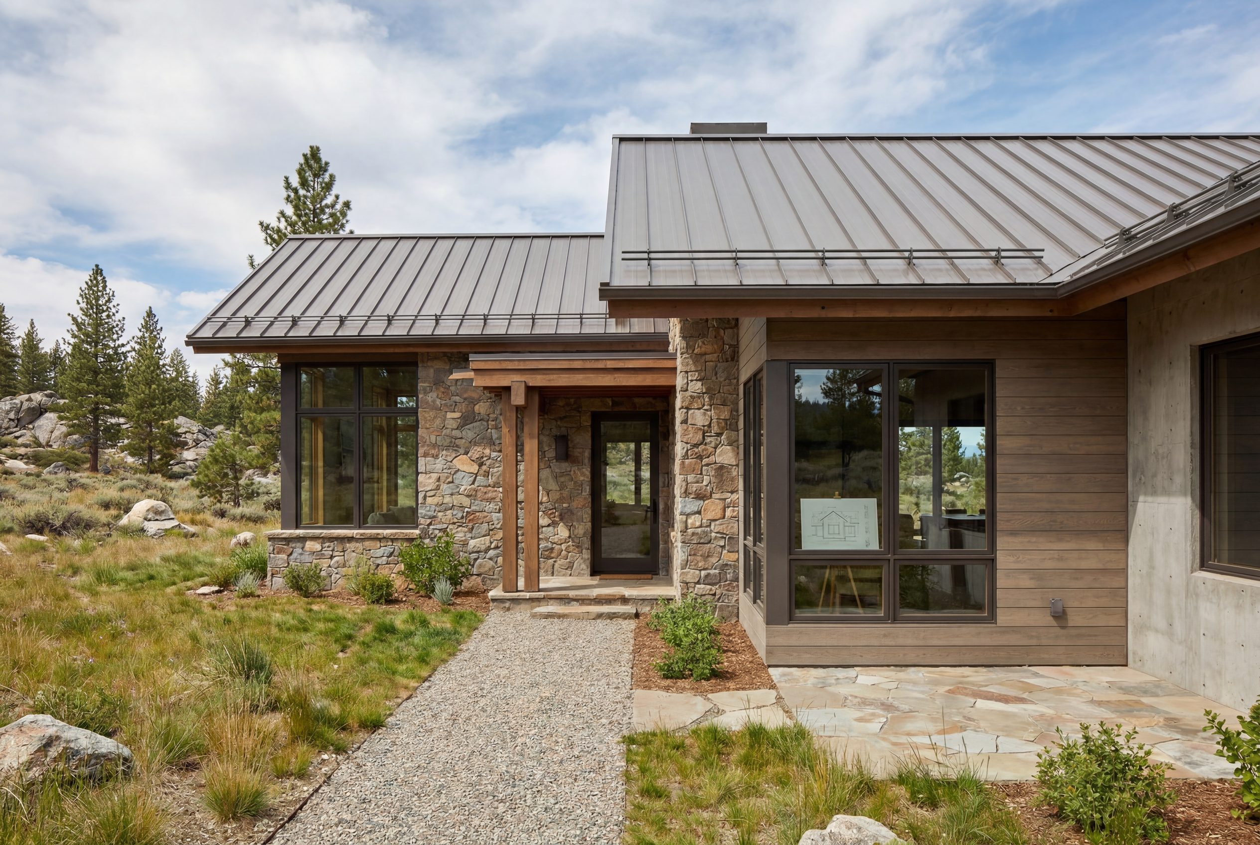 Entry approach of a contemporary ranch home &ndash; stone masonry, weathered wood siding, and standing-seam metal roofs among granite and pines